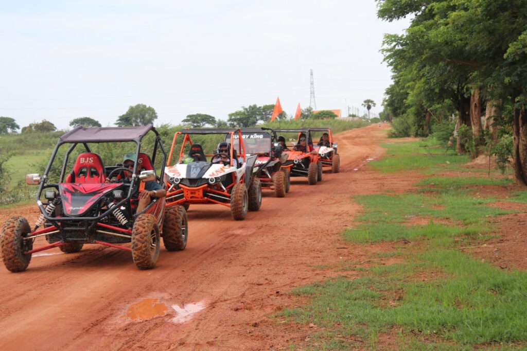 A line of five off-road buggies in different colors driving down a wide red dirt road lined with green grass and trees under a clear sky.