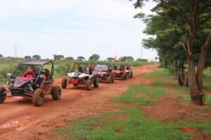 A line of five colorful off-road buggies driving along a rural red dirt road, led by a driver in a red and black buggy, with green trees and fields in the background.