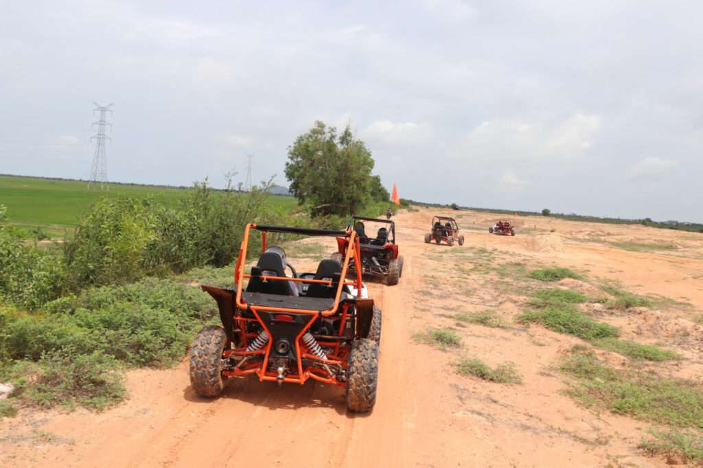 A line of orange and black off-road buggies driving along a dirt path through open fields under a bright sky.