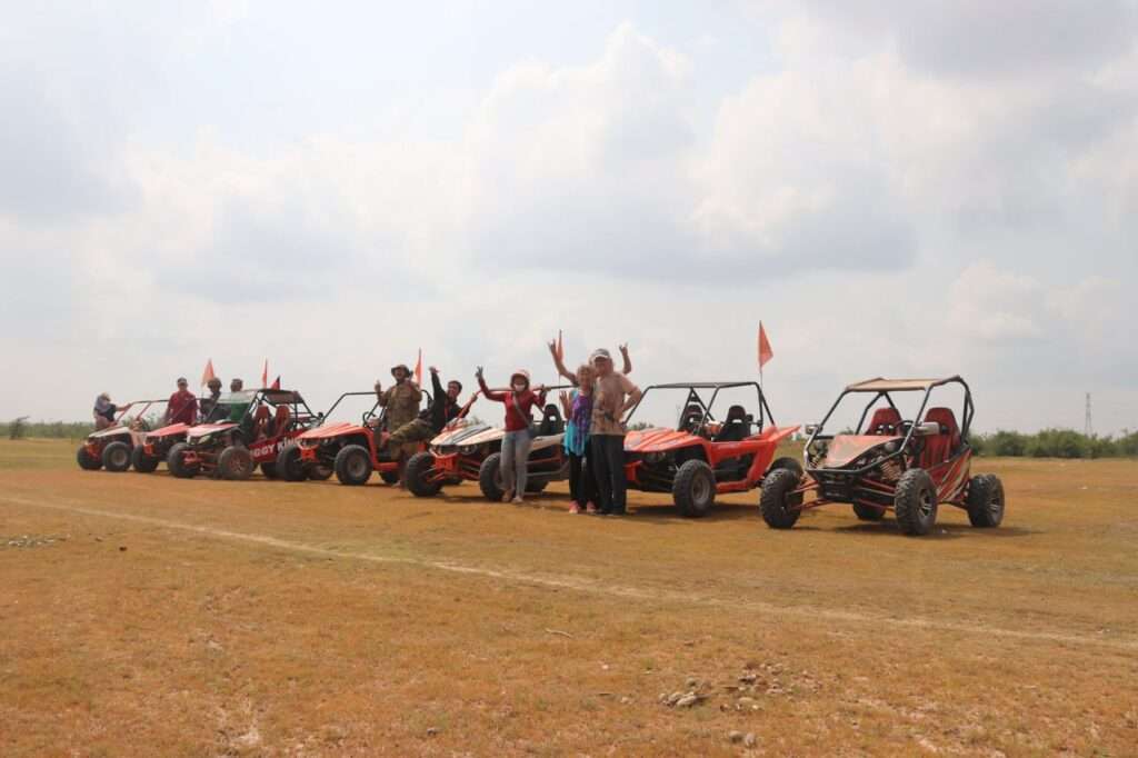 A group of tourists and guides posing cheerfully between a row of orange and red off-road buggies in a wide, dry field under a cloudy sky.