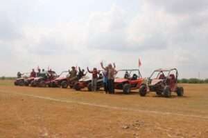 A group of tourists and guides posing cheerfully between a row of orange and red off-road buggies in a wide, dry field under a cloudy sky.