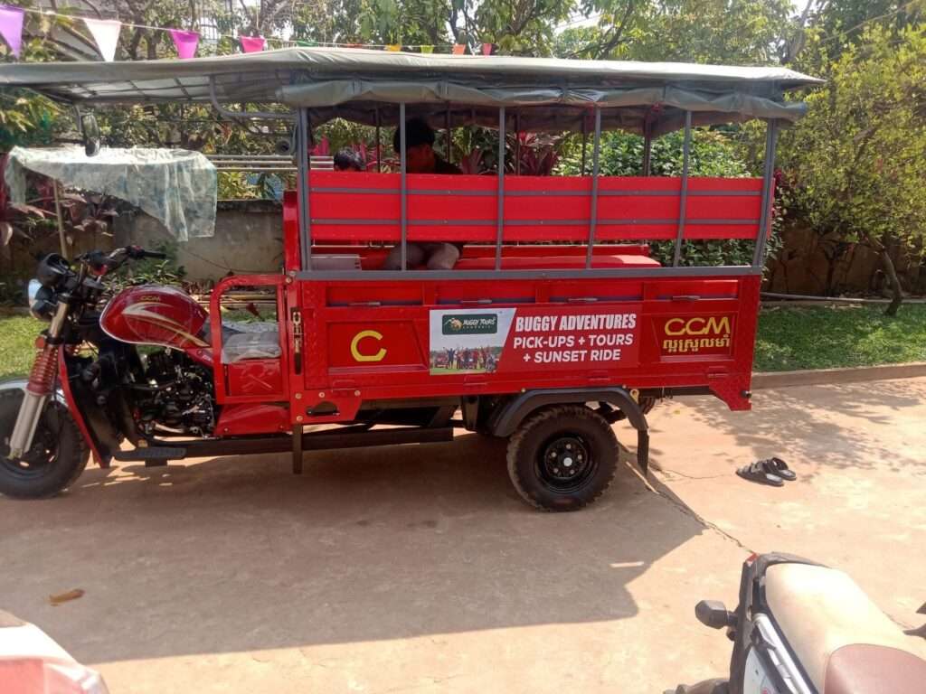 A bright red passenger motorized tuk-tuk vehicle with a canopy roof, featuring a side sign that reads 'Buggy Adventures Pick-Ups + Tours + Sunset Ride' parked on a paved driveway.