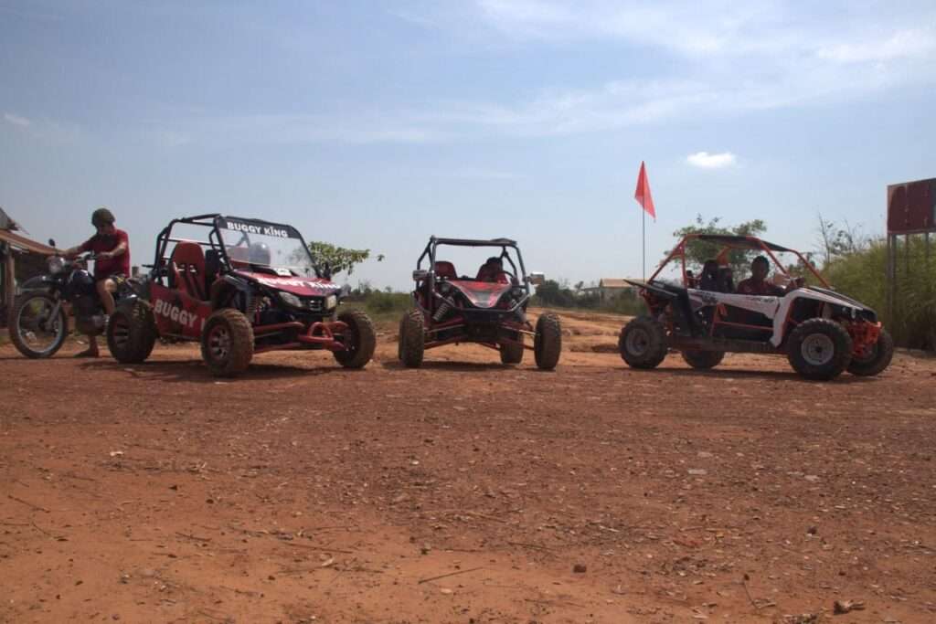 Three off-road adventure buggies lined up on a red dirt trail next to a tour guide on a motorcycle under a blue sky.