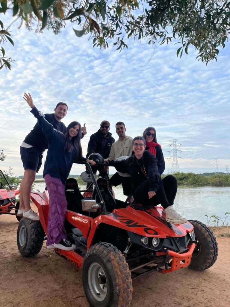 A group of six smiling friends posing on and around a bright orange off-road adventure buggy parked near a river under a cloudy sky.