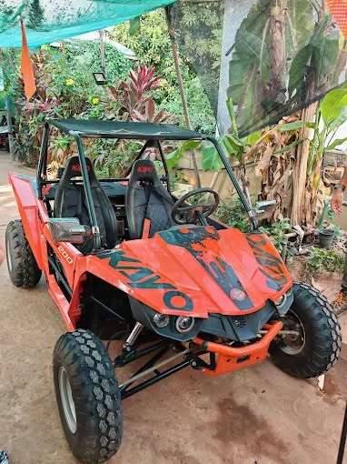 A close-up shot of a bright orange Kayo S200 two-seater off-road buggy with large treaded tires, black bucket seats, and a roll cage, parked in a tropical outdoor setting.