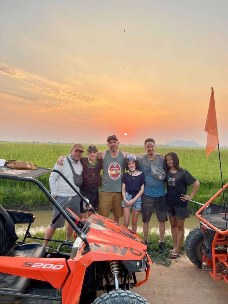 A group of five smiling tourists standing together between two orange off-road buggies in front of a green rice field during a vibrant golden sunset.