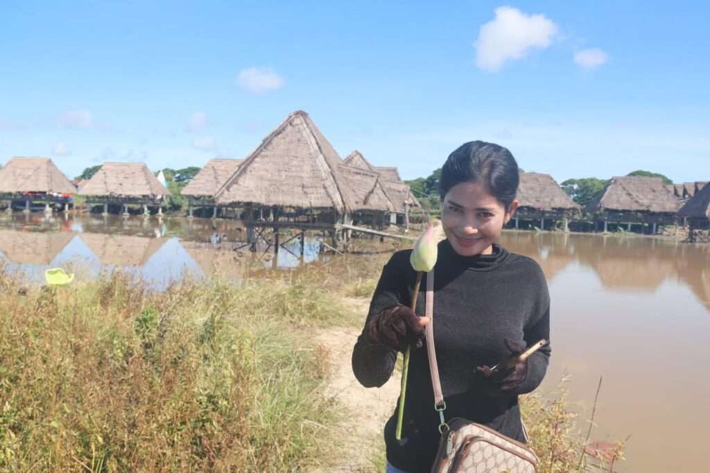 A smiling woman in a black shirt holding a lotus flower bud stands near a body of water with thatched-roof stilt huts in the background.