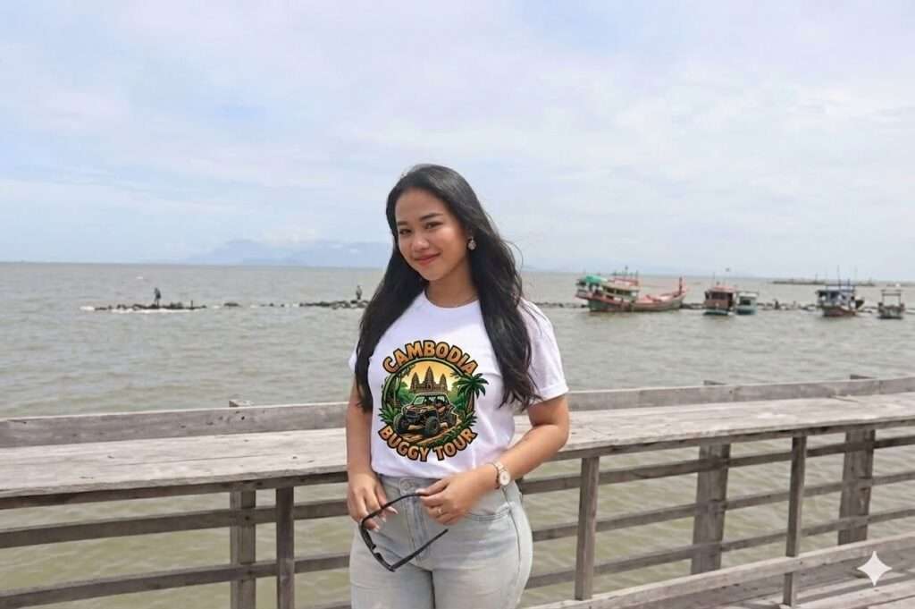 A smiling woman standing on a wooden pier by the water, wearing a white Cambodia Buggy Tour t-shirt and holding sunglasses, with boats visible in the background.