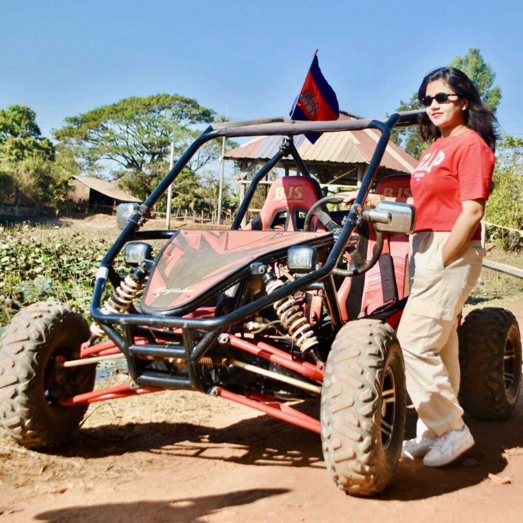 A female tourist wearing sunglasses and a red shirt stands next to a black and red off-road buggy flying a Cambodian flag on a rural dirt path.