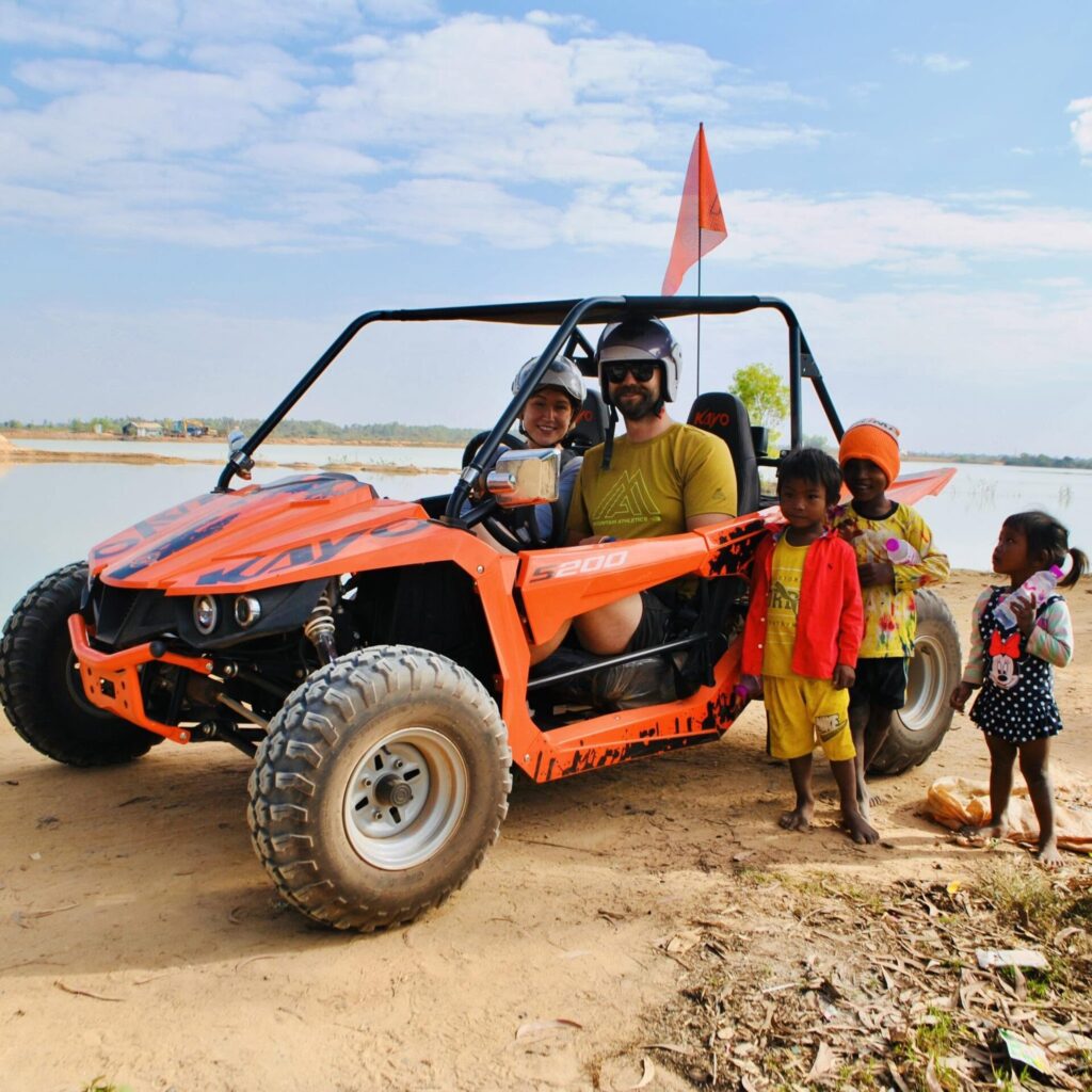 A man and woman wearing helmets sit inside an orange off-road buggy next to local Cambodian children smiling by a lakeside.