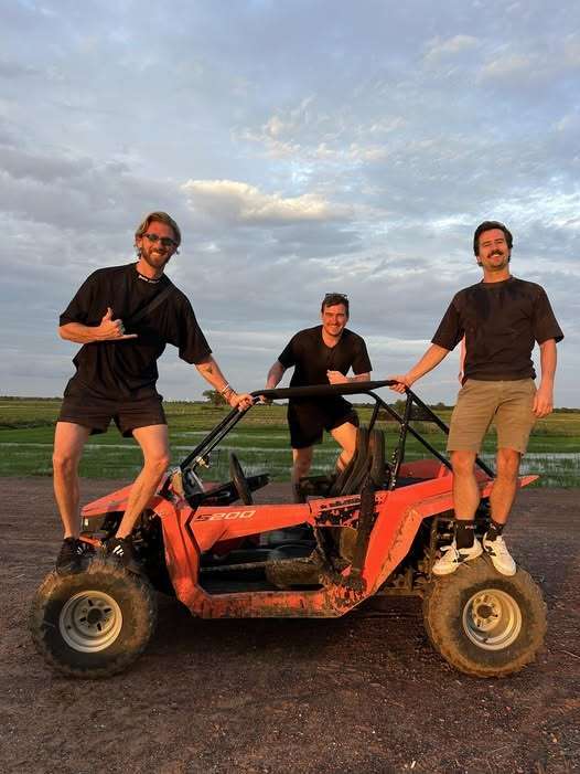 Three smiling male tourists posing on and around an orange off-road buggy on a dirt trail in Cambodia at sunset.