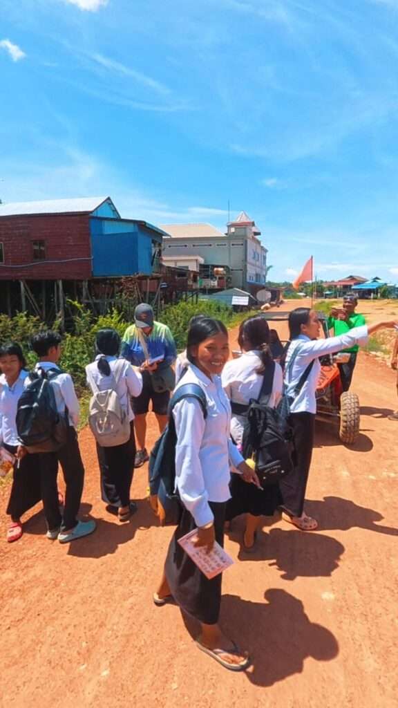 Cambodian students in white school uniforms walking along a dirt road past stilt houses during a rural buggy tour stop near Siem Reap.
