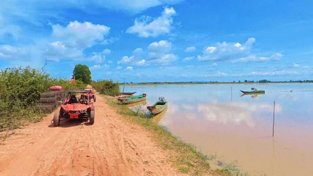 Red off-road buggy driving along a dirt path beside Tonle Sap Lake with traditional Cambodian wooden boats moored nearby under a blue sky.