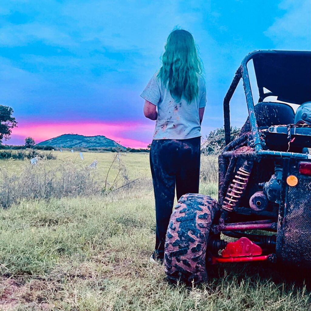 A person with turquoise-tinted hair and mud-spattered clothes stands facing away from the camera in a grassy field next to an off-road buggy, gazing at a vibrant pink sky hovering over distant hills at dusk